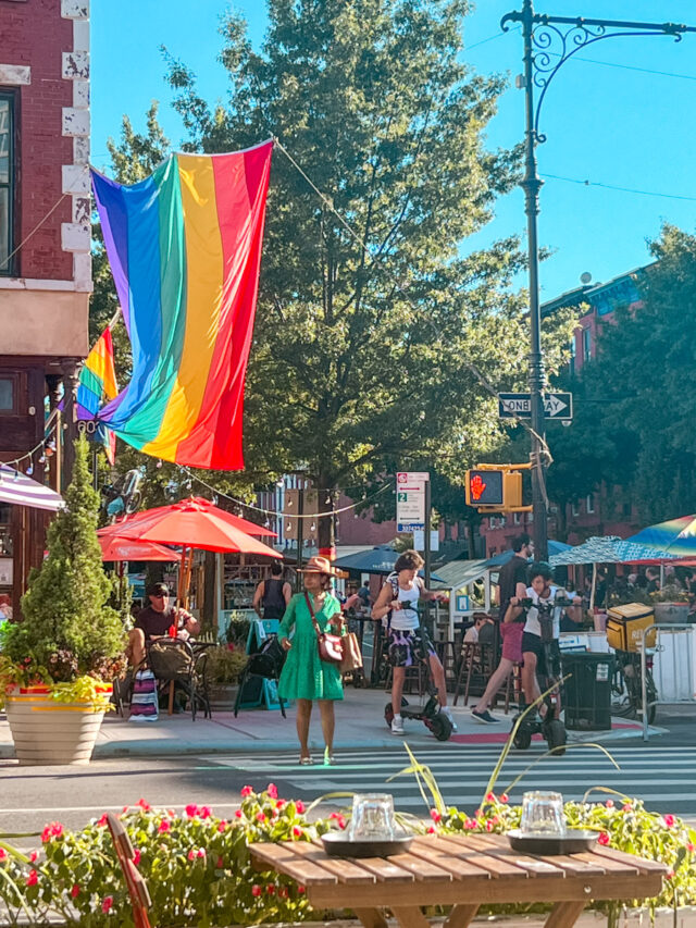 People crossing a street in a queer NYC neighborhood decorated with rainbow flags.