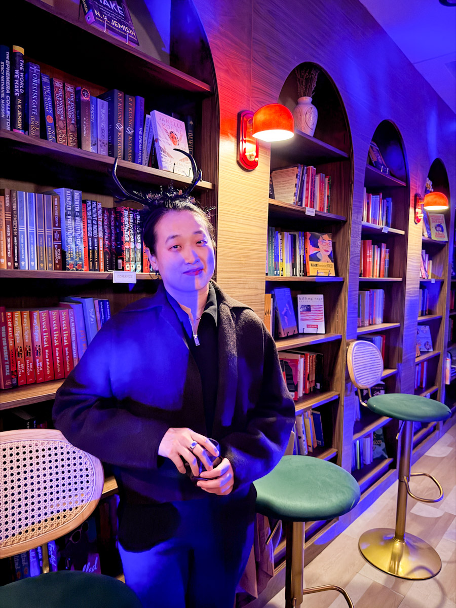 A woman in black Halloween costume standing in front of bookshelves inside a NYC bookstore and wine bar.