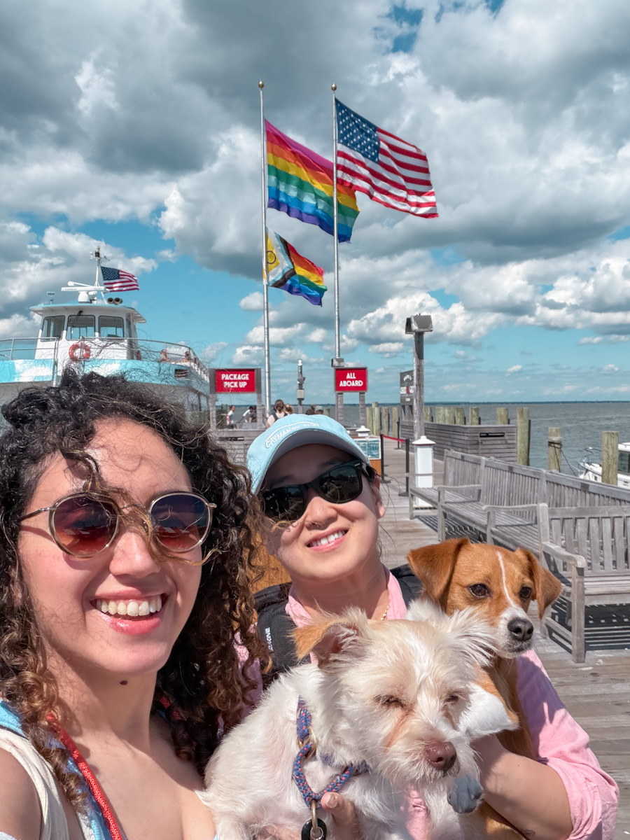 Two women carrying dogs on a ferry dock in Cherry Grove, Fire Island in New York with a ferry and US and queer flags on the background.
