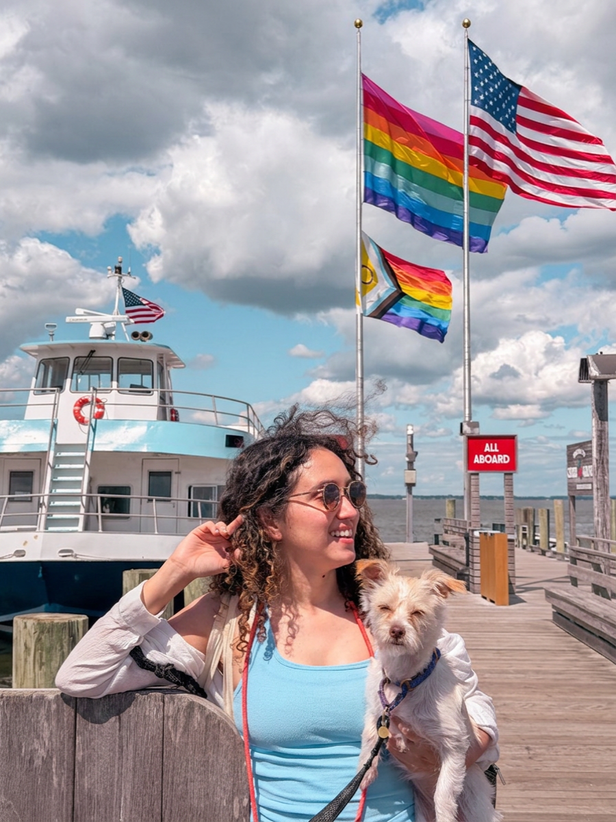 A woman holding a dog on a ferry dock in Cherry Grove, Fire Island, New York with a ferry and USA and queer flags in the background.