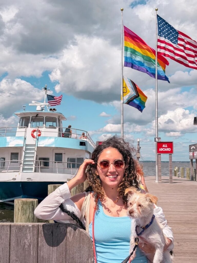 A woman wearing a pair od sunglasses and holding a dog on a ferry dock in Cherry Grove, Fire Island, New York with a ferry and USA and queer flags in the background.
